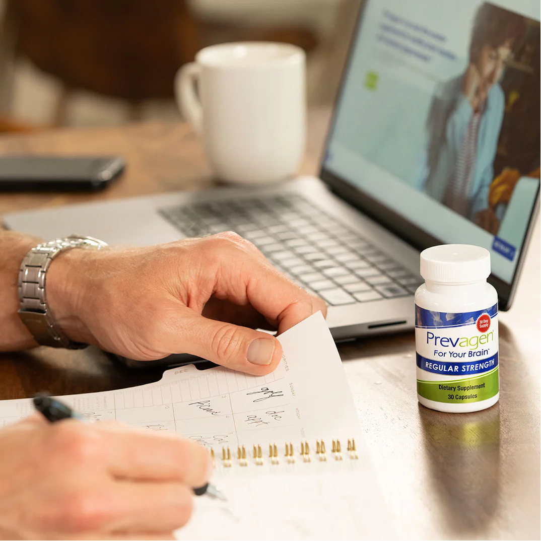 Person writing in a notebook with a bottle of Prevagen on a desk next to a laptop.